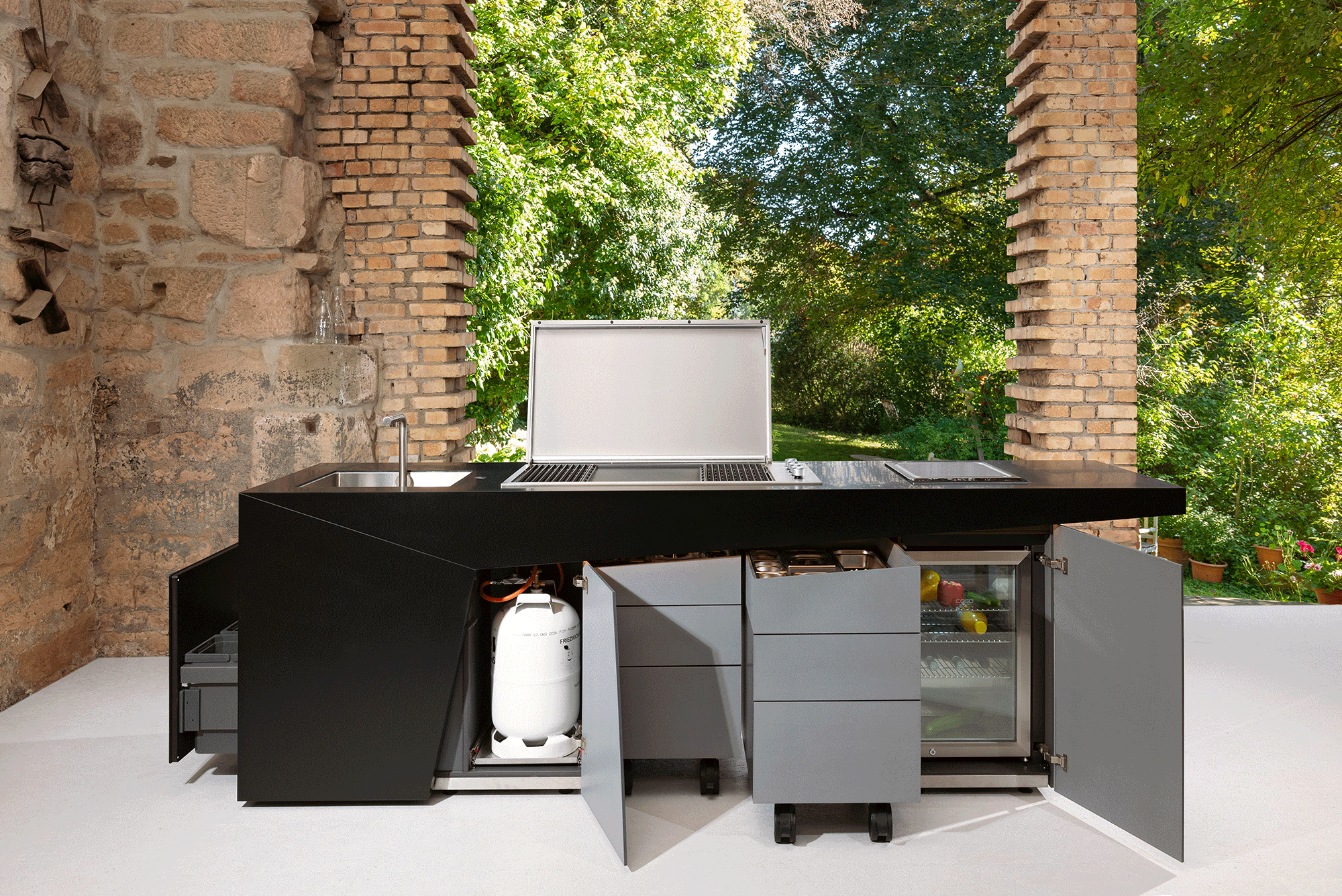 Küchenzeile mit geöffneten Fronten in einem zum Garten hin offenen Raum mit Mauerwerk aus Naturstein und Backsteinen