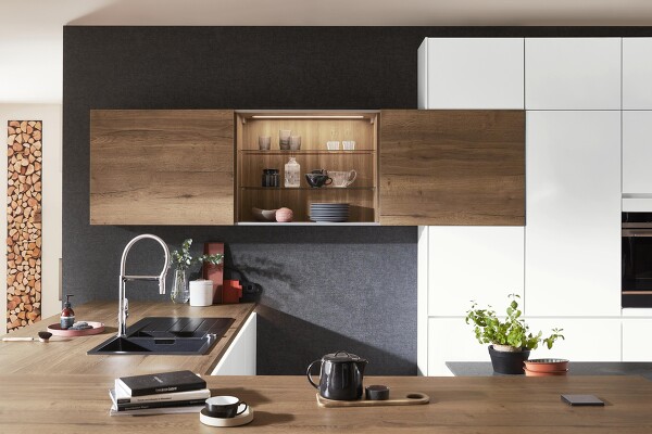 Perfektes Zusammenspiel von Farben und Licht. Die matte alpinweiße Küche und die dunkle Wand fassen Arbeitsplatte und Schränke aus Holzoptik optimal ein. Tolles Extra: Wandschrank mit Schiebetüren, der durch seine Innenbeleuchtung mehr Behaglichkeit bringt.